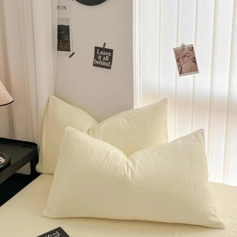 A set of minimalist solid color duvet cover and pillowcases in a cream or off-white shade on a bed.
