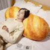 A woman rests her head on a large, golden-yellow plush baguette shaped pillow while wearing a cream sweater.