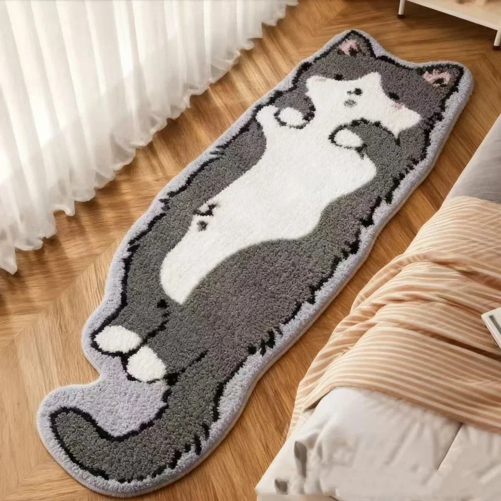 A gray and white cartoon cat plush rug lying on a wooden floor next to a bed with a beige blanket.