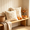 A beige throw pillow cover rests on a wooden bench next to a woven basket and dried floral arrangements.