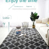 A dark gray geometric plush area rug is displayed on a light wooden floor in a bright living room setting.