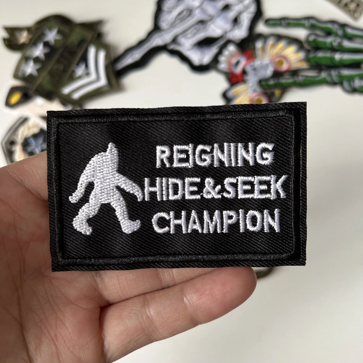 A black embroidered patch with white text that reads Reigning Hide&Seek Champion.
