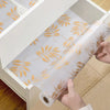 A roll of white silicone non-slip drawer liner with gold leaf pattern being pulled out of a white storage box.