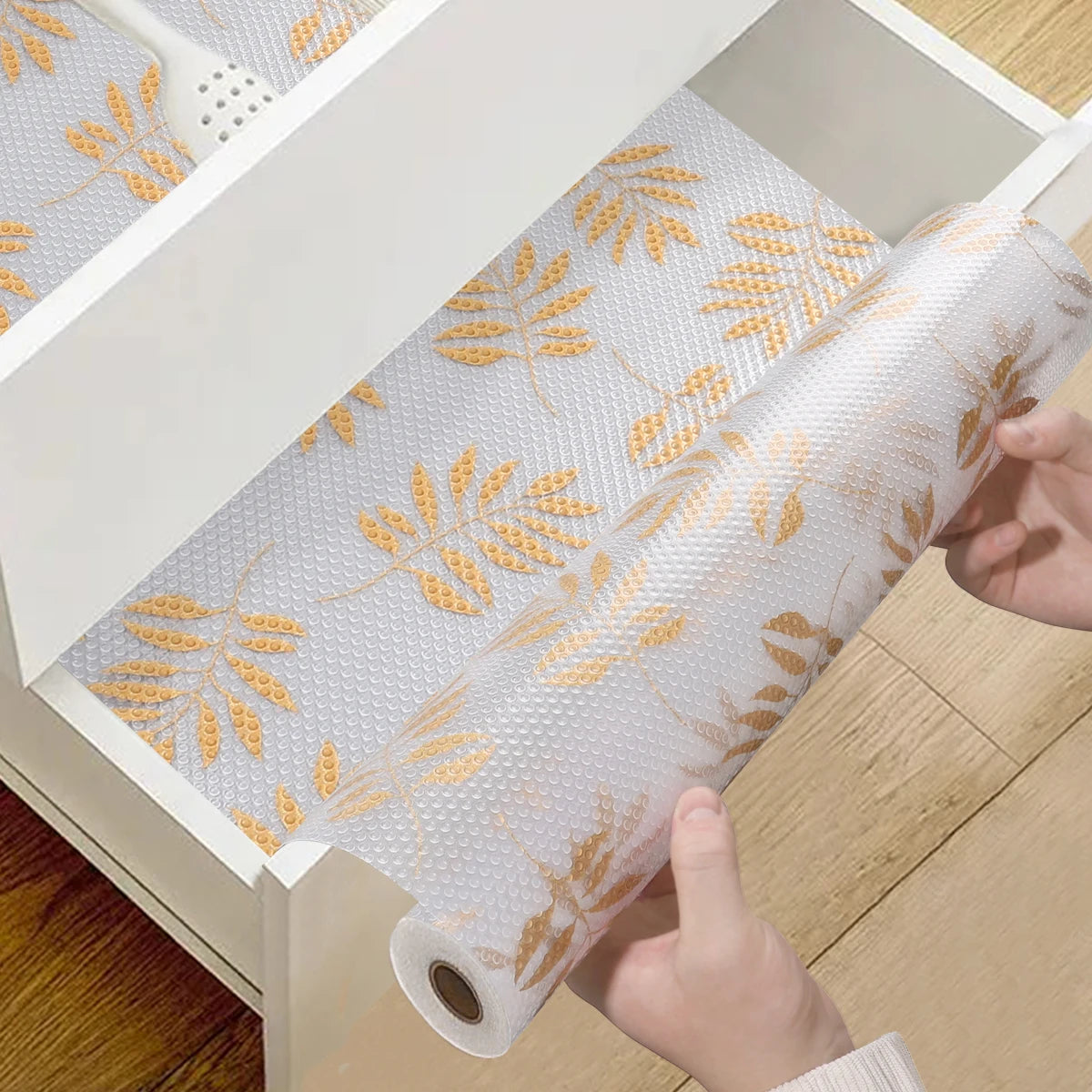 A roll of white silicone non-slip drawer liner with gold leaf pattern being pulled out of a white storage box.