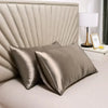 A set of two luxurious silk pillow covers in a champagne or taupe color resting on a bed.