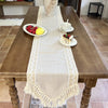 A wooden table is set with a beige macrame lace table runner, fruit, and a small dessert.