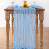 Light blue cheesecloth table runner draped over a wooden table with yellow sunflowers and glassware.