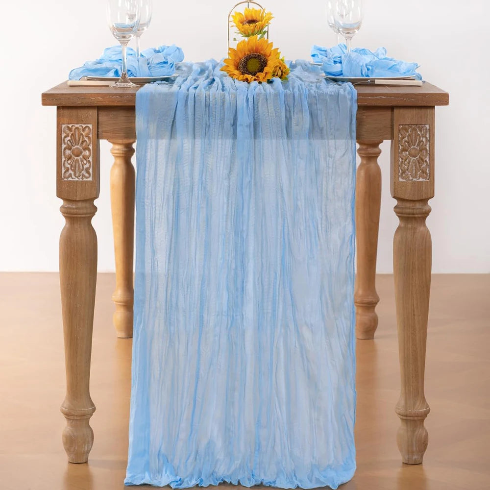 Light blue cheesecloth table runner draped over a wooden table with yellow sunflowers and glassware.