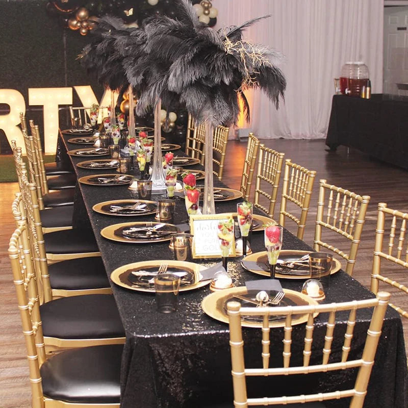 A long table set for an event featuring a black sequin glitter tablecloth, gold Chiavari chairs, and floral centerpieces.