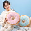 A young woman holds a pink frosted doughnut plush pillow with sprinkles and a blue doughnut plush pillow in a light blue sett