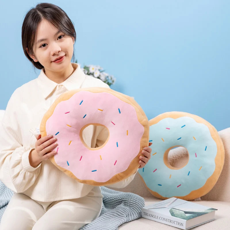 A young woman holds a pink frosted doughnut plush pillow with sprinkles and a blue doughnut plush pillow in a light blue sett