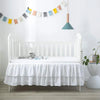 A white wooden crib with a ruffled skirt and a hanging light fixture in a bright nursery room.