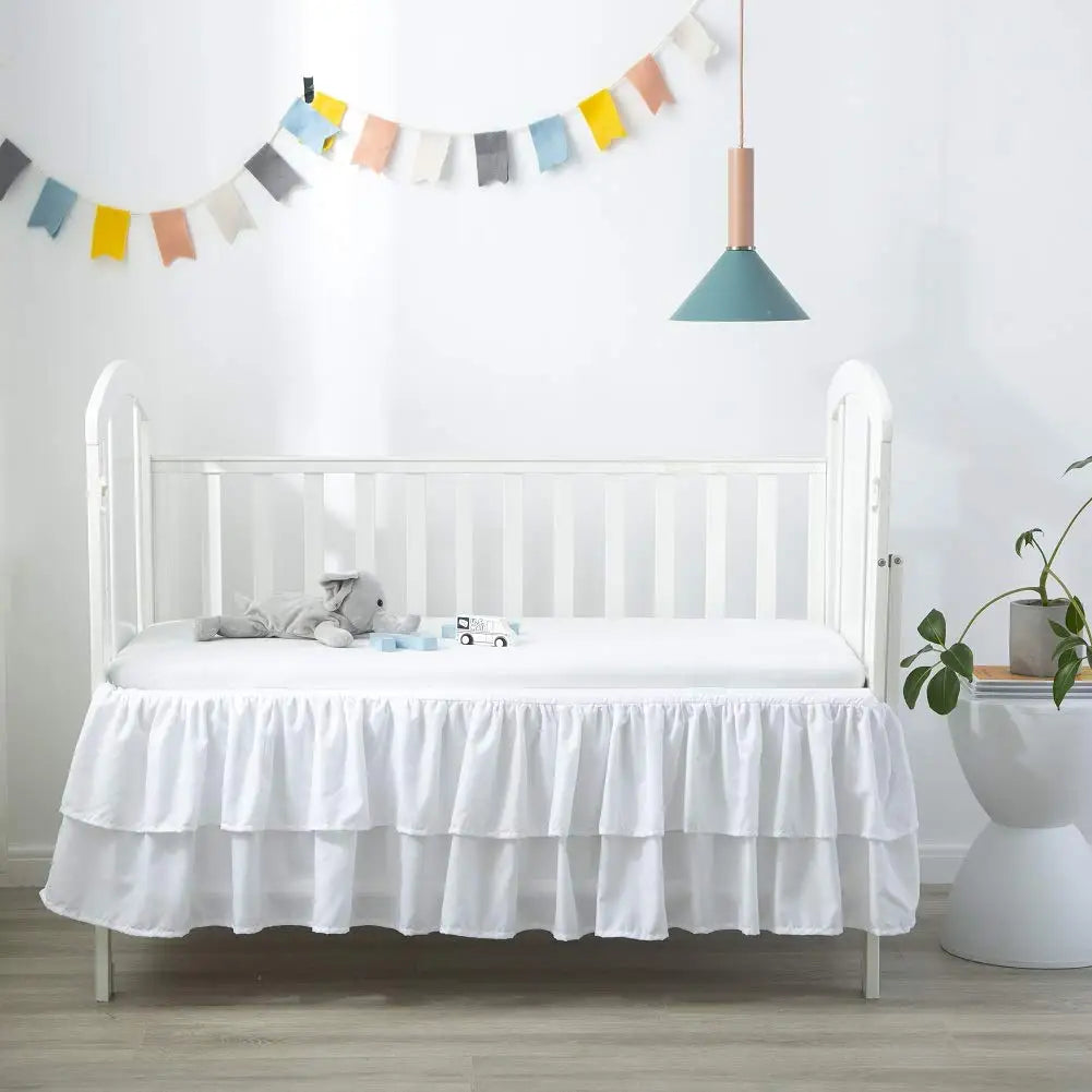 A white wooden crib with a ruffled skirt and a hanging light fixture in a bright nursery room.