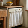 A white kitchen curtain skirt with ruffle trim and ribbon ties hanging on a dishwasher rack.