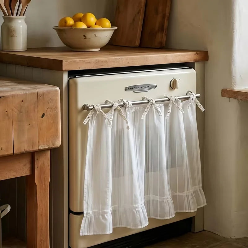 A white kitchen curtain skirt with ruffle trim and ribbon ties hanging on a dishwasher rack.