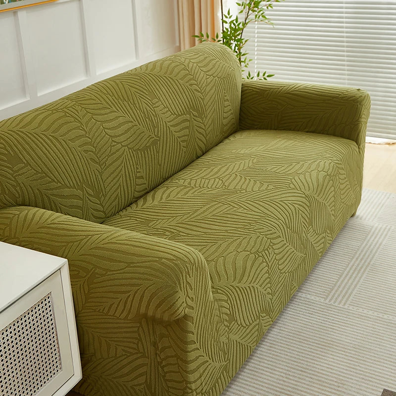 A moss green sofa slipcover featuring a jacquard leaves pattern, shown in a bright interior setting.