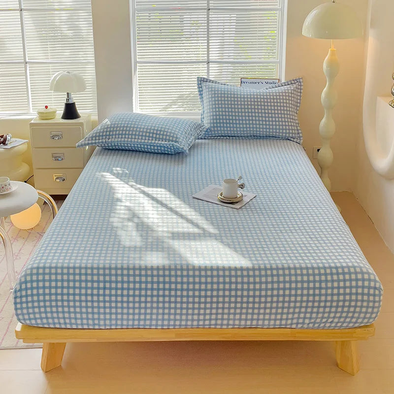 A light blue checkered flannel fitted sheet is laid out on a wooden bed frame in a bright bedroom.