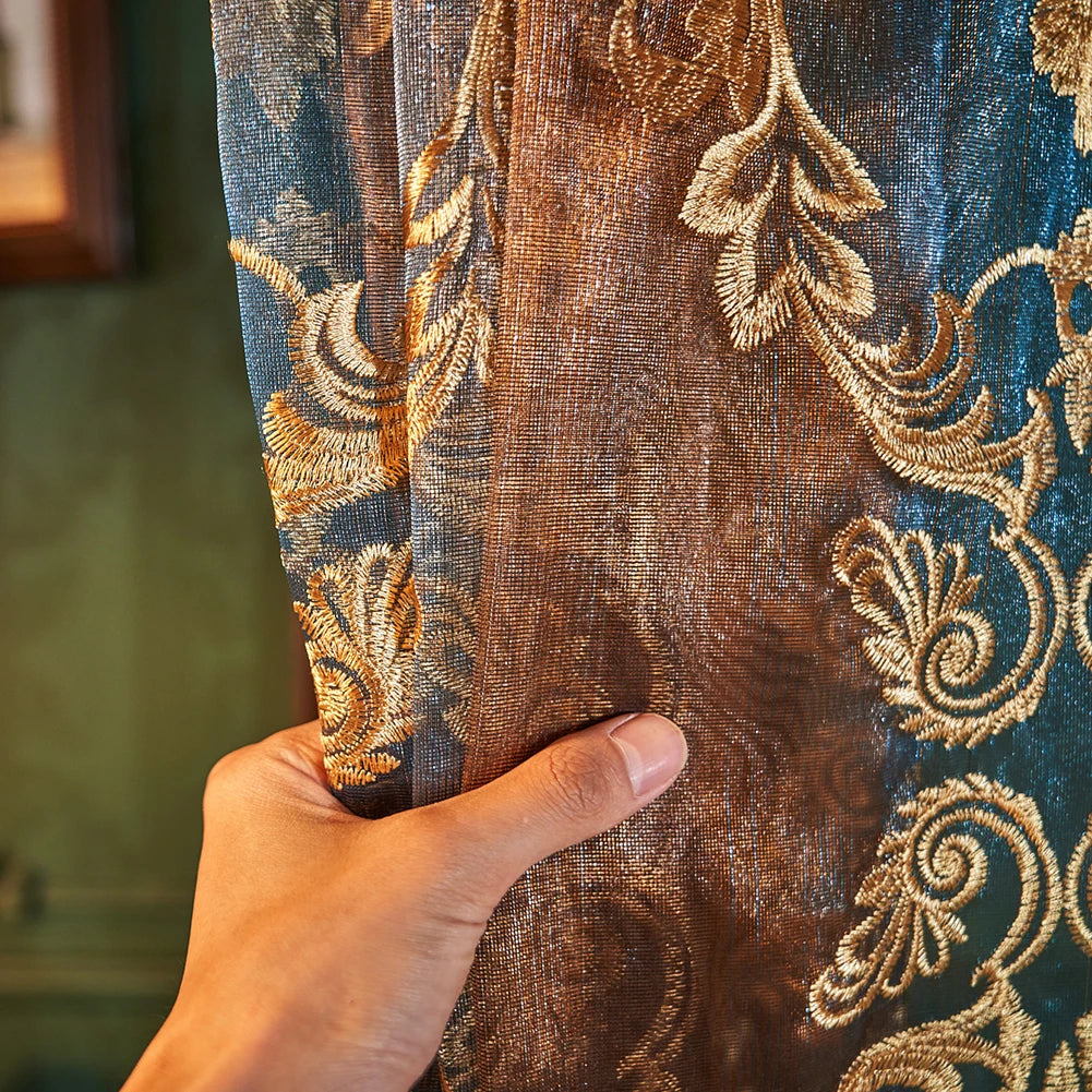 A hand touches a close-up of brown sheer curtains with gold floral embroidery.