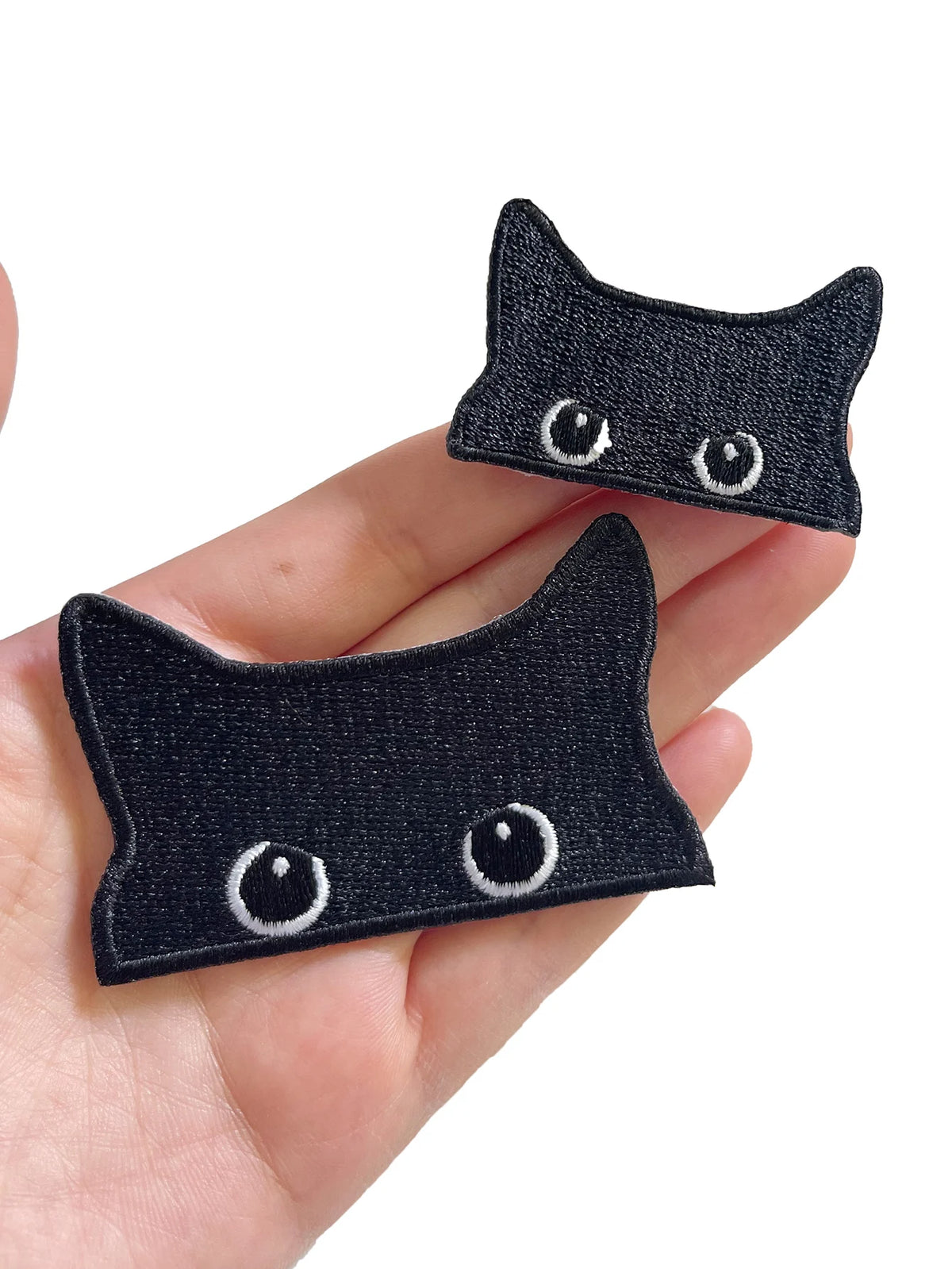A pair of embroidered black cat iron-on patches with white eyes are shown being held in a hand.