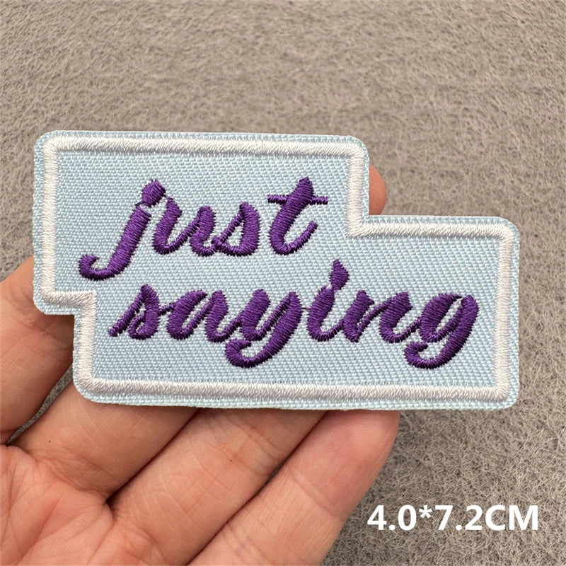 A light blue embroidered patch with the purple text Just saying is held in a hand.