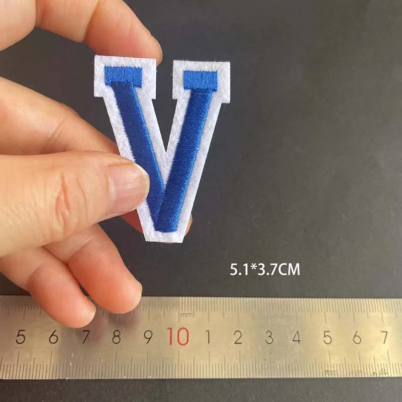 A hand holds a blue embroidered letter V patch against a ruler showing its size.