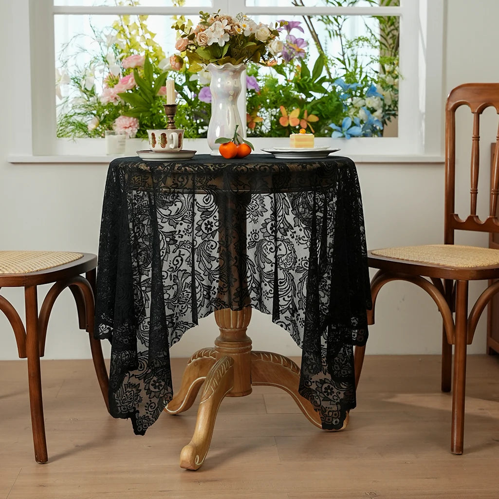 A black lace tablecloth is draped over a wooden table, set for dining with flowers visible in the background.