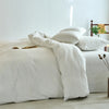 A white cotton yarn-dyed duvet cover is displayed on a bed with natural light and a wooden shelf in the background.