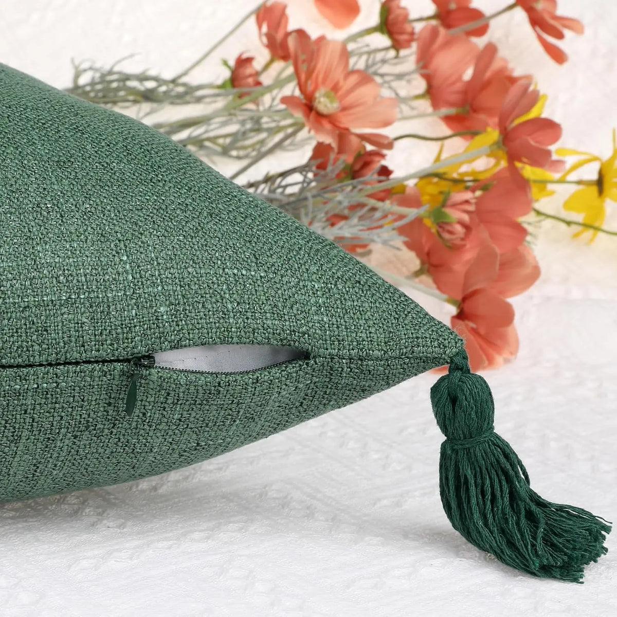 A dark green boho cotton linen throw pillow cover with a tassel, resting on a white surface next to dried flowers.