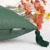 A dark green boho cotton linen throw pillow cover with a tassel, resting on a white surface next to dried flowers.