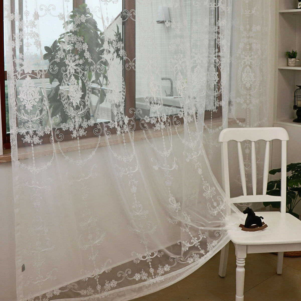 White sheer curtains with floral embroidery draped over a glass door and next to a white wooden chair.