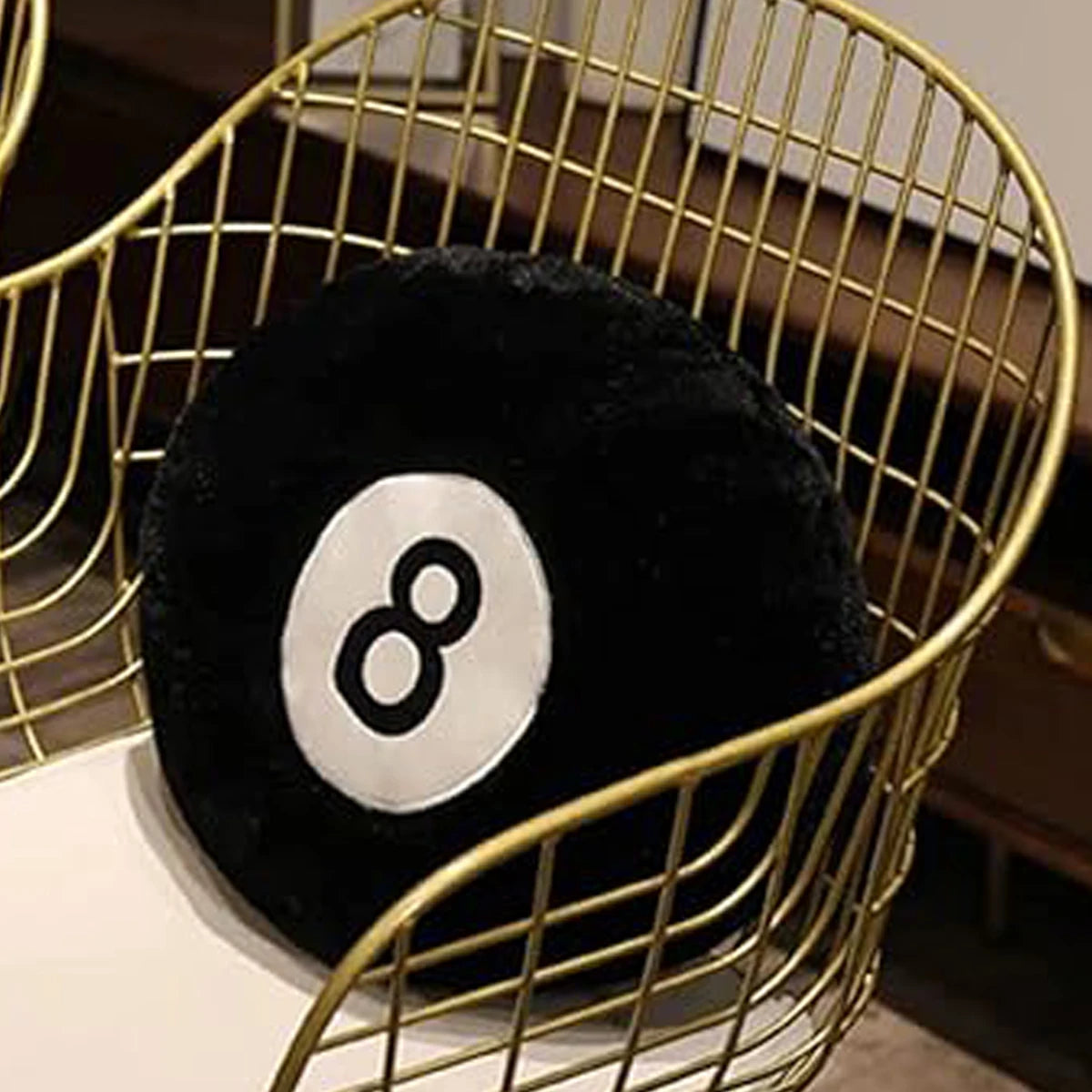 A black memory foam ball pillow cushion with a white number eight design is displayed in a gold wire basket.