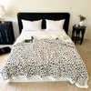 A white leopard print knit throw blanket is draped over a bed with white pillows.