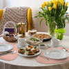 A white table set with pink placemats, plates, silverware, and food, surrounded by yellow and beige textiles.
