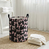 A black and pink patterned foldable canvas laundry basket sitting on a woven mat next to white towels.