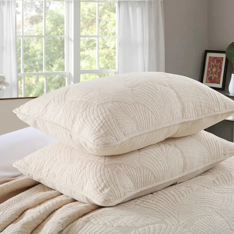 A set of cream embroidered cotton quilted pillows resting on a beige quilted bedspread near a window.