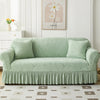 A light sage green jacquard elastic sofa cover with a pleated skirt displayed on a white wooden cabinet.