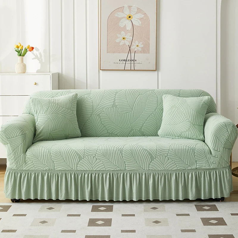 A light sage green jacquard elastic sofa cover with a pleated skirt displayed on a white wooden cabinet.