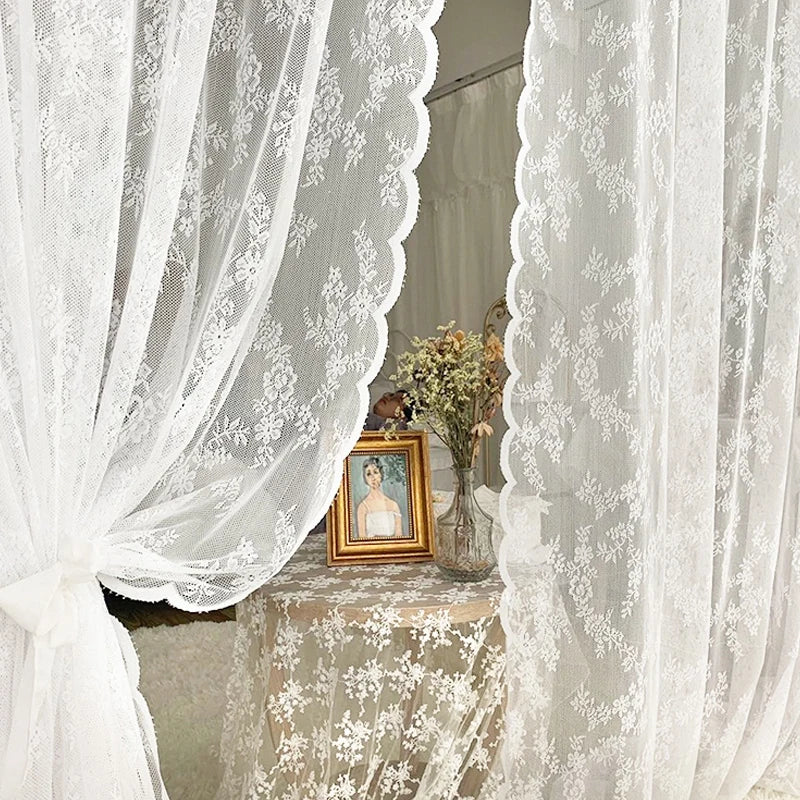 White sheer floral lace window curtains framing a display of flowers and a framed portrait.