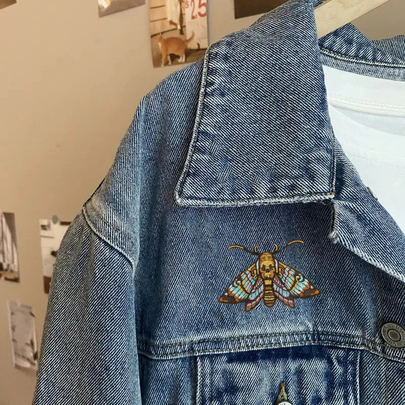 A close-up of a denim jacket featuring an embroidered colorful moth patch on the chest.