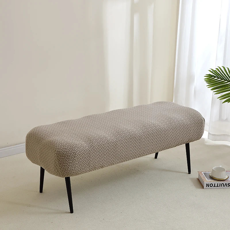 A beige elastic jacquard bench cover shown on a light floor, with a window and a green plant in the background.