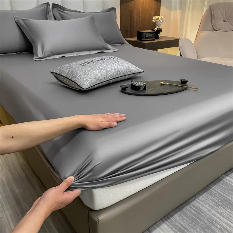 A close-up of a gray fitted bed sheet draped over a bed, with pillows and a watch on the sheet.