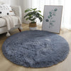 A round, fluffy gray shaggy area rug placed on a light wooden floor next to a plant and framed artwork.