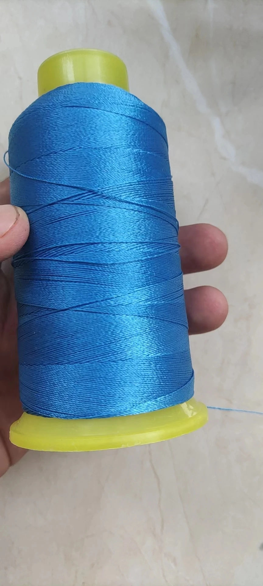 A spool of bright blue high tenacity polyester sewing thread is held in a hand, with a yellow plastic cap visible.