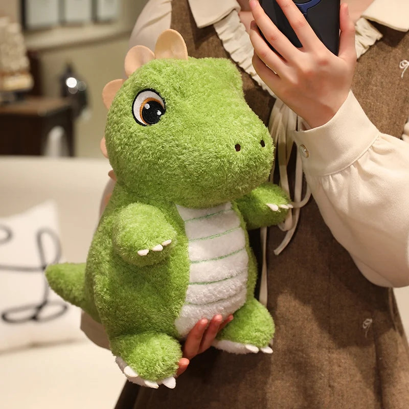 A hand holds a cute green dinosaur plush toy with white accents against a brown garment.