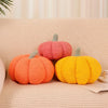 Three knitted pumpkin shaped throw pillows in shades of orange, pink, and yellow on a light beige sofa.
