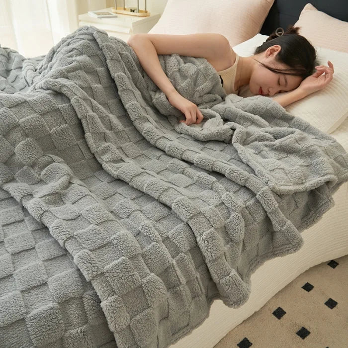 A woman is relaxing under a cozy gray checkered fleece throw blanket on a bed.