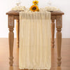 A beige cheesecloth table runner draped over a wooden table with floral accents for boho wedding decor.