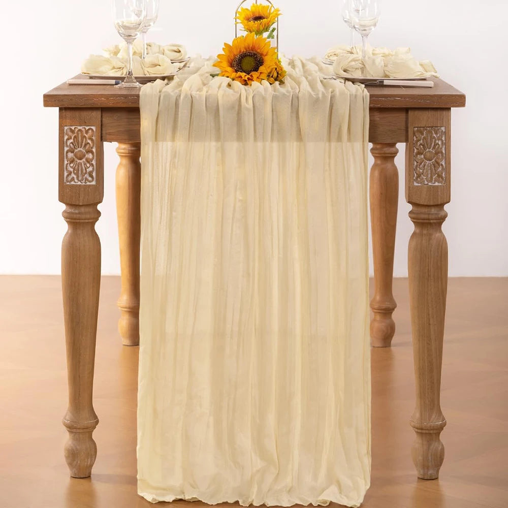 A beige cheesecloth table runner draped over a wooden table with floral accents for boho wedding decor.