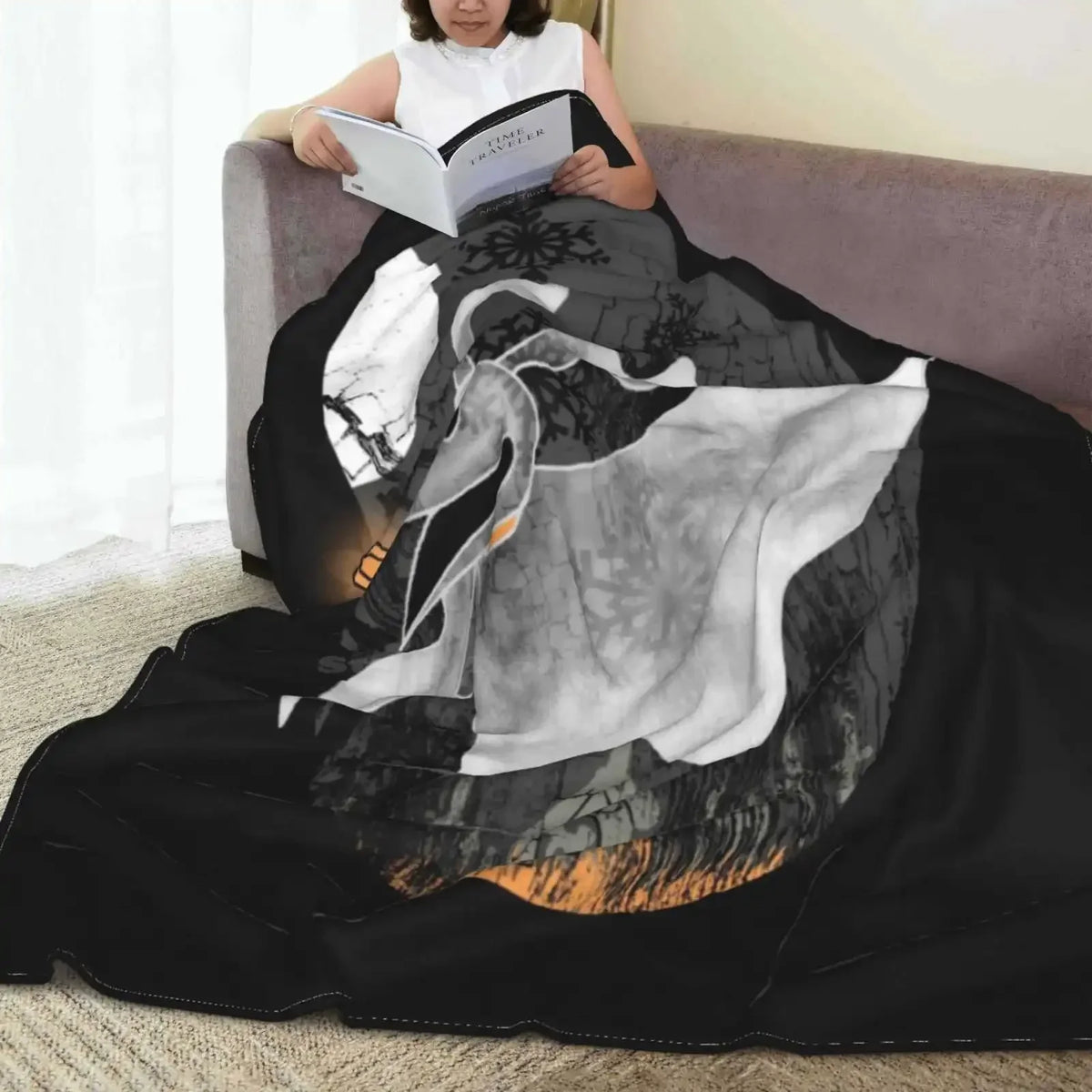 A person is sitting on a couch reading a book while a black and white patterned microfiber throw blanket is draped over the s