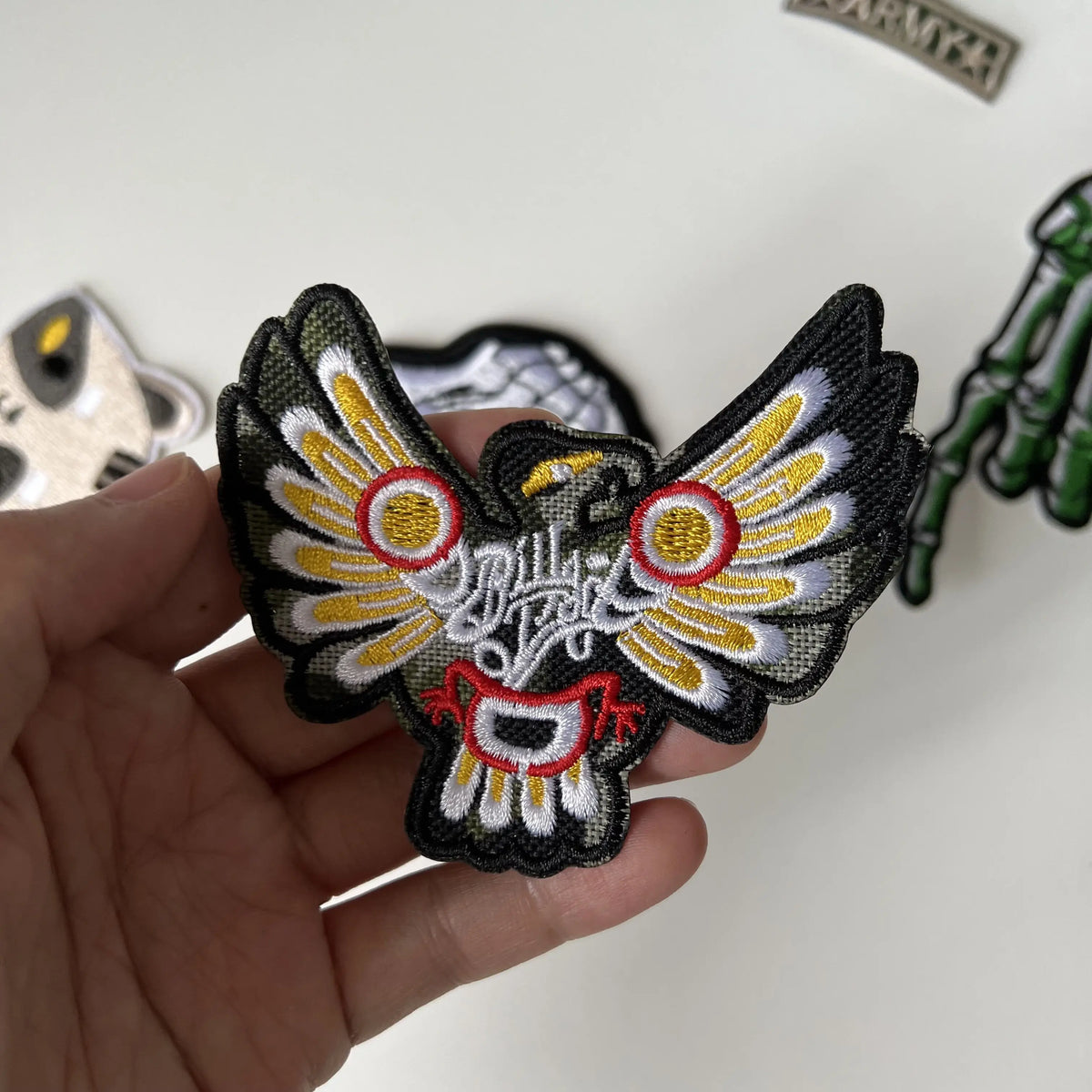 A hand holds a black embroidered patch featuring a stylized eagle with yellow and white feathers and red accents.